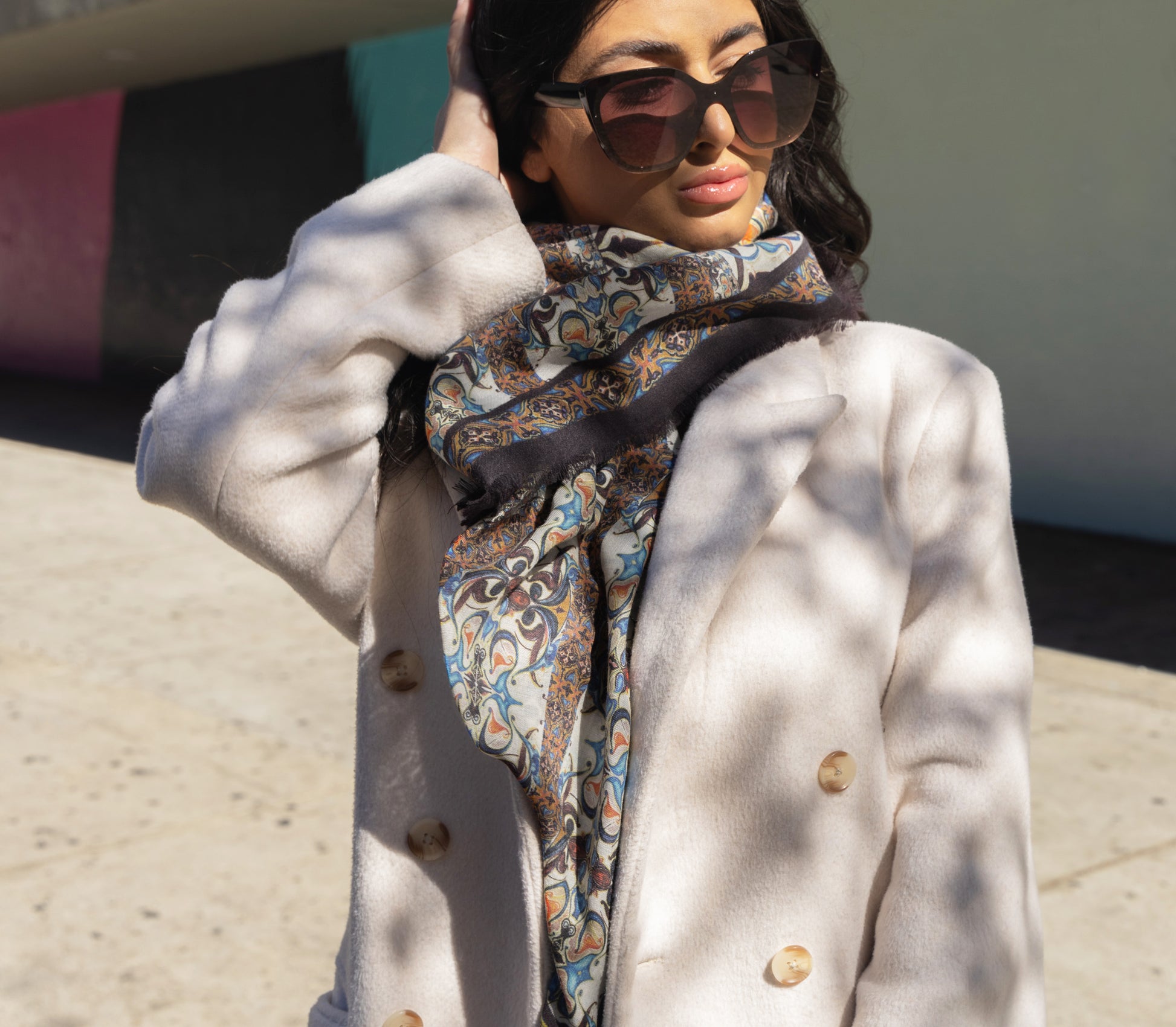 A woman wearing sunglasses and a floral scarf with intricate patterns, in shades of yellow, burgundy, and blue, wrapped around her neck.