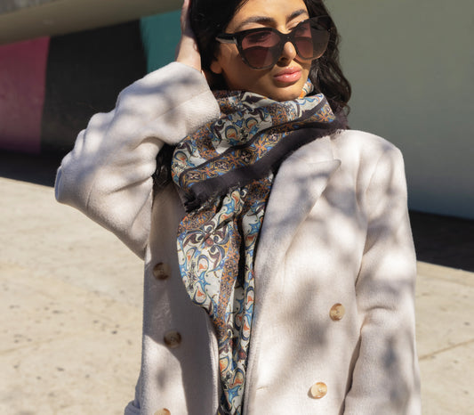 A woman wearing sunglasses and a floral scarf with intricate patterns, in shades of yellow, burgundy, and blue, wrapped around her neck.