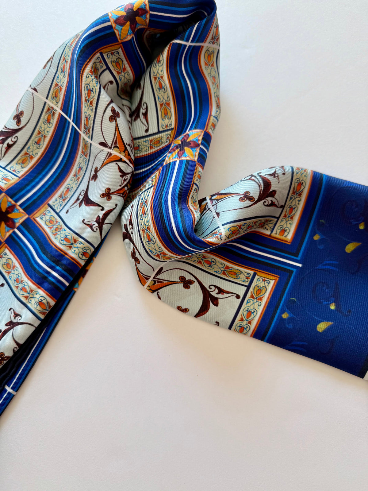 A folded silk scarf with a traditional Mediterranean pattern featuring blue, white, brown, and gold colors, displayed on a clean white background.